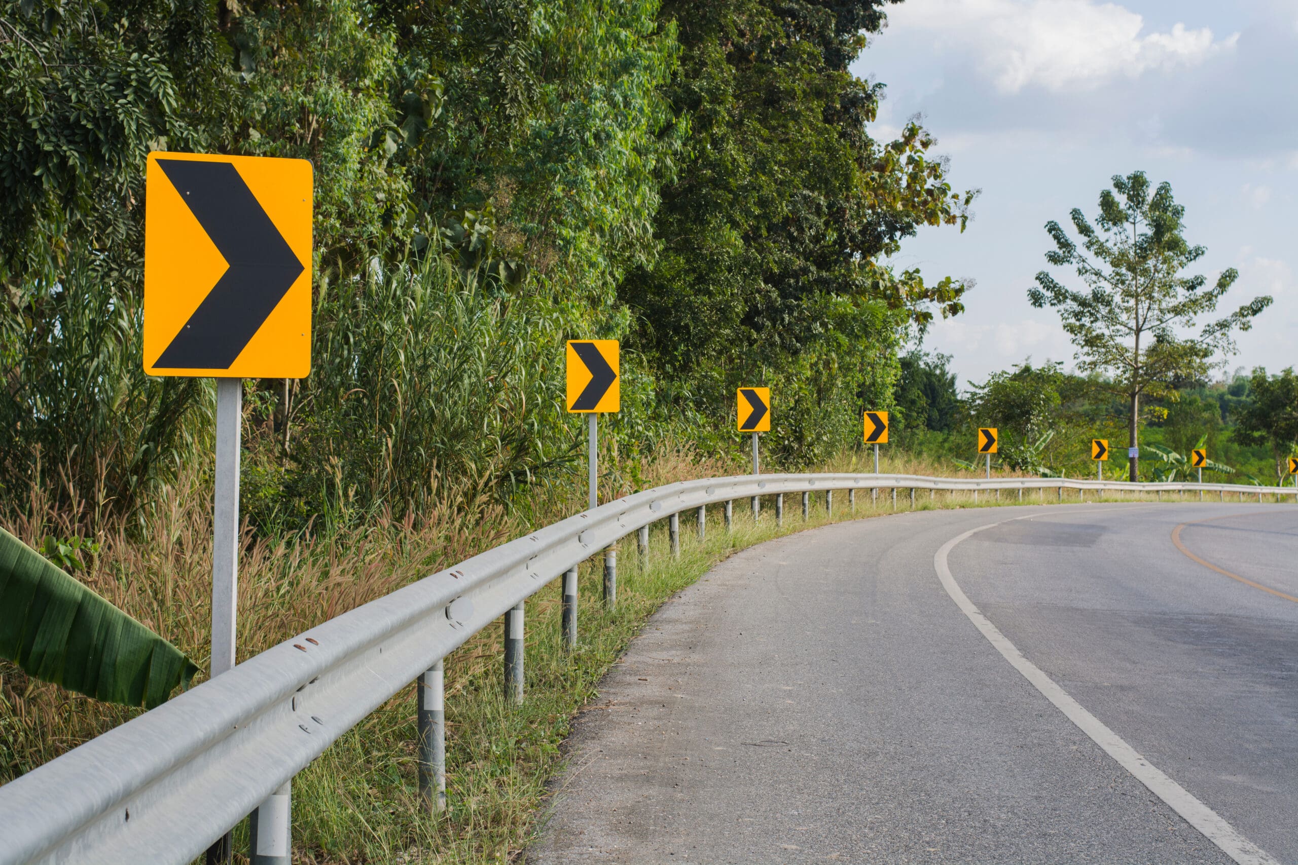 Chevron signs positioned behind a guard rail as seen from the perspective of a driver approaching a curve, with trees and sky in the background.